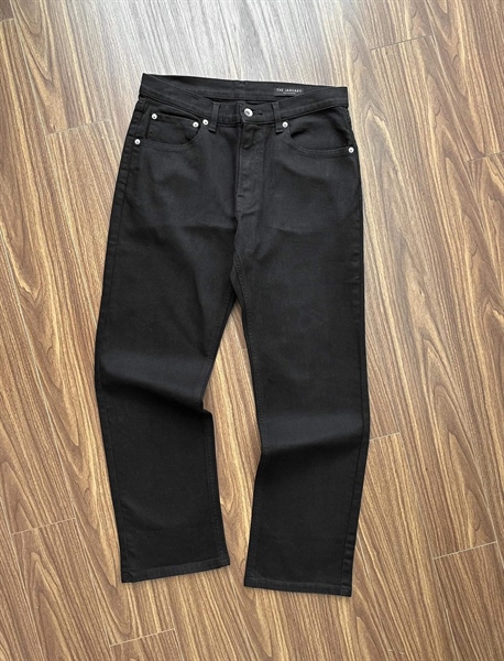 Quần jeans đen dáng ống đứng trải phẳng mặt trước trên sàn gỗ, cạp có nút và đinh tán kim loại