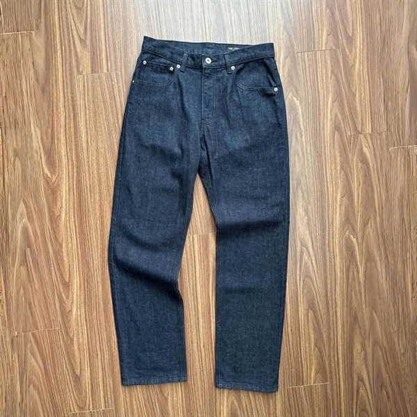 Quần jeans xanh chàm đậm dáng ống đứng trải phẳng mặt trước trên sàn gỗ, túi trước và nút kim loại