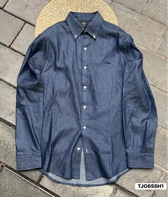 Áo sơ mi dài tay màu xanh denim đậm, cổ cài nút, logo thêu nhỏ ở ngực, trải phẳng trên nền gạch