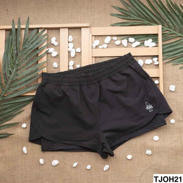Quần short nữ đen hai lớp, cạp chun, gấu bo tròn, trải phẳng trên nền bao bố với lá cọ và sỏi trắng