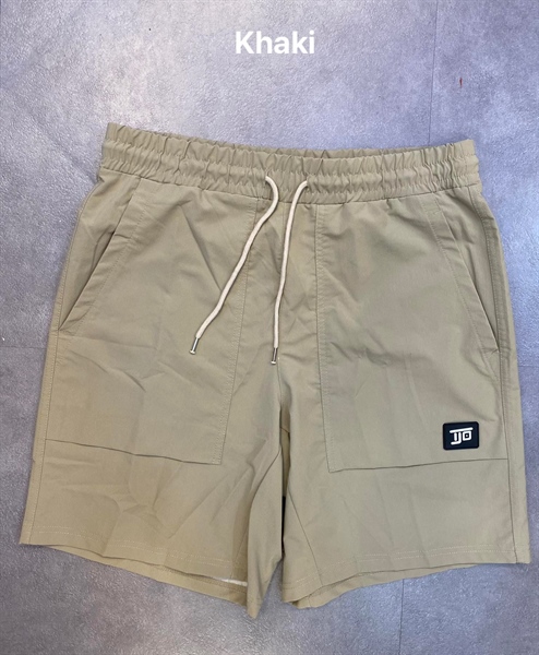 Quần short màu kaki lưng thun dây rút kem, túi đắp hai bên, nhãn logo đen nhỏ, trải phẳng trên nền xám