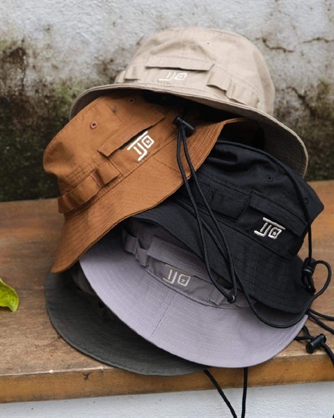 Chồng mũ bucket nhiều màu be, nâu bò, đen, xám, xanh rêu xếp trên ghế gỗ, có dây rút và logo thêu